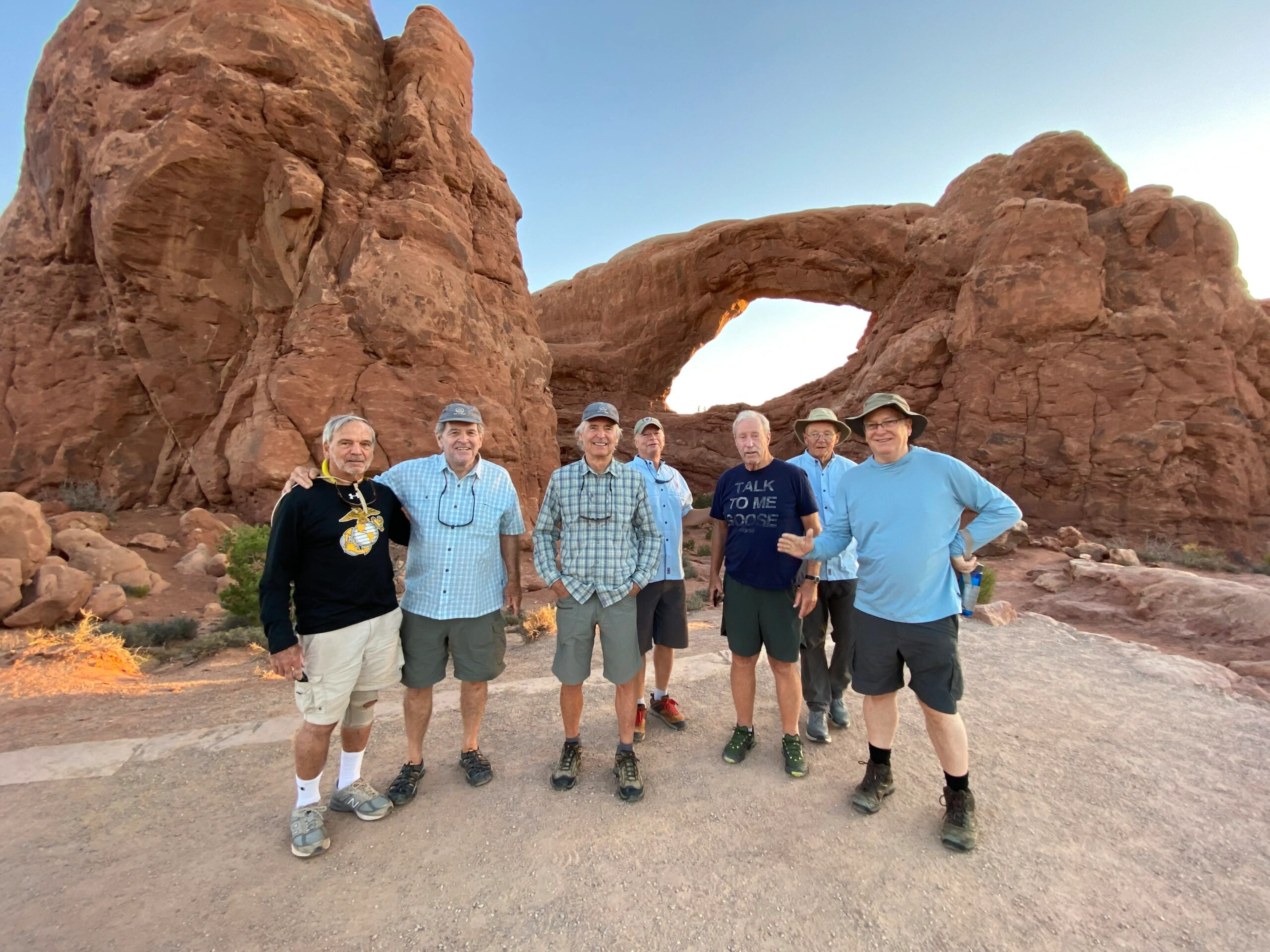 Seven men stand in front of a large natural sandstone arch in a rocky desert landscape, with clear sky in the background.