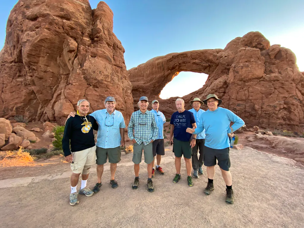 Seven men pose in front of a large rock arch formation in a desert landscape, wearing casual outdoor clothing and hats.