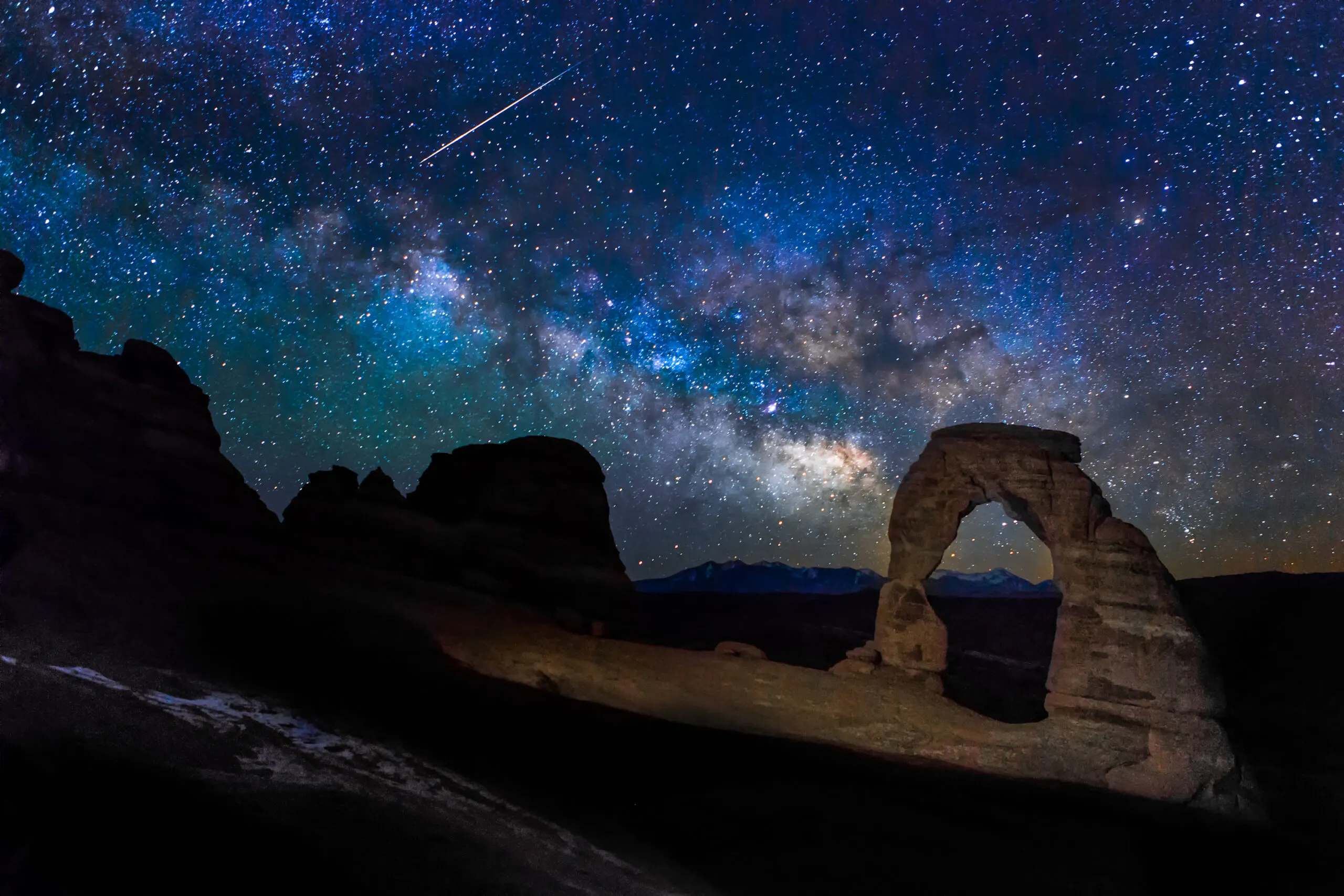 A star-filled night sky with a visible shooting star above Delicate Arch rock formation in Arches National Park.