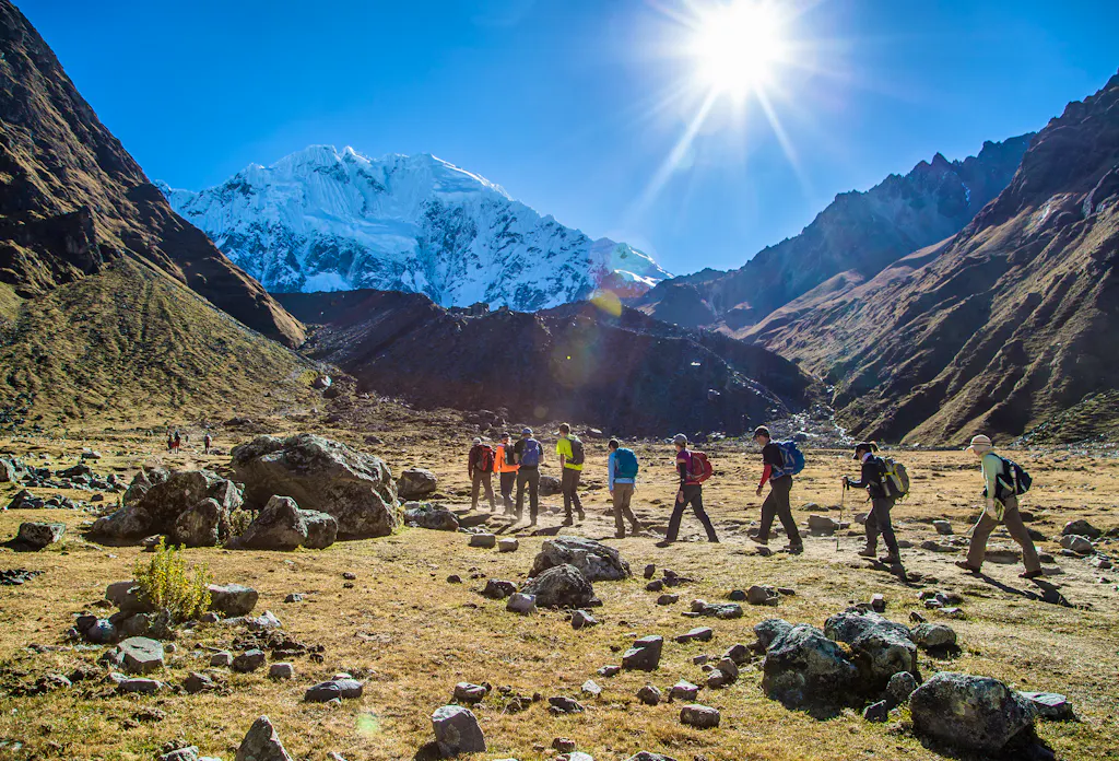 A group of hikers with backpacks walk through a rocky mountain valley under a bright sun, with a snow-covered peak in the background.