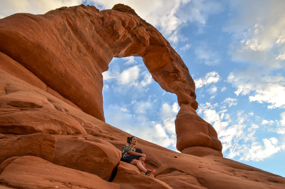 A person sits on a rock beneath Delicate Arch, a large natural sandstone arch, with a partly cloudy sky in the background.