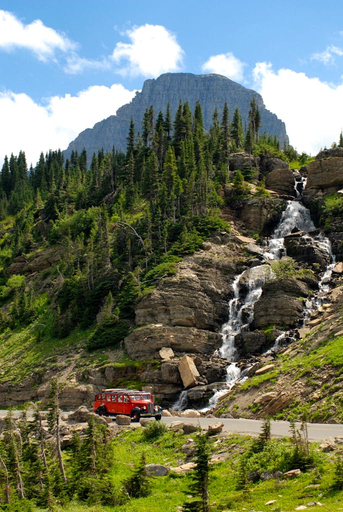 A red tour bus passes by a waterfall at Logan Pass on Glacier Park's Going to the Sun Highway