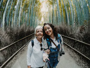 Two women stand on a path through a dense bamboo forest, smiling and taking a selfie together. Both wear backpacks filled with travel resources; one holds a selfie stick, and a camera hangs from her neck.