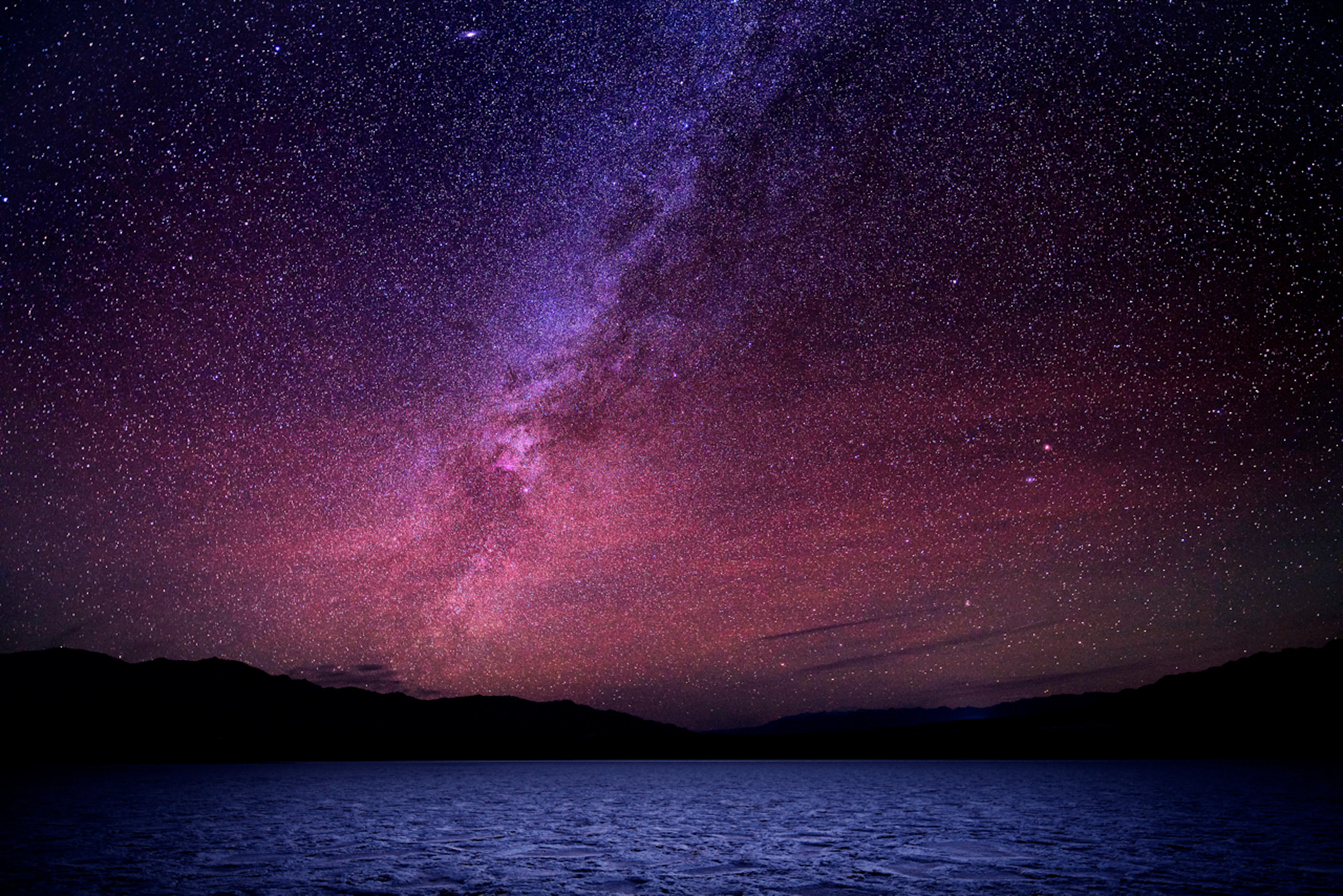 A night sky filled with stars and the Milky Way galaxy above a silhouette of mountains and a calm body of water—an unforgettable scene from one of the best hikes in North America.