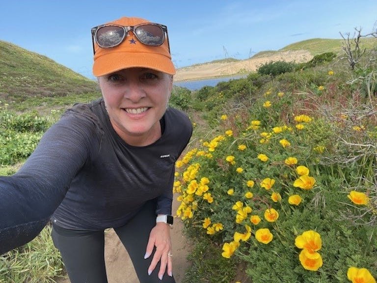 Person wearing an orange cap and sunglasses, crouching beside yellow flowers on a hiking trail in a hilly, grassy area under a clear sky near Point Reyes National Seashore in North California