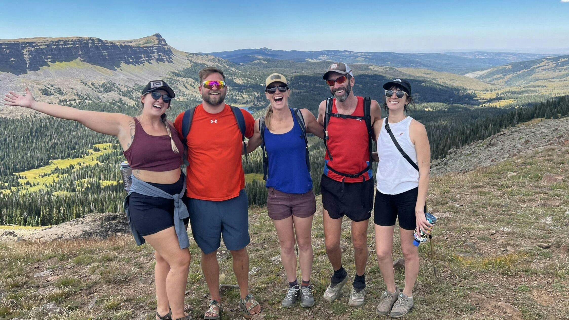 Five people standing on a mountain trail with a scenic view of valleys, hills, and a clear blue sky in the background. Some are wearing sunglasses and hats, and a few are smiling with arms around each other in Denver, Colorado
