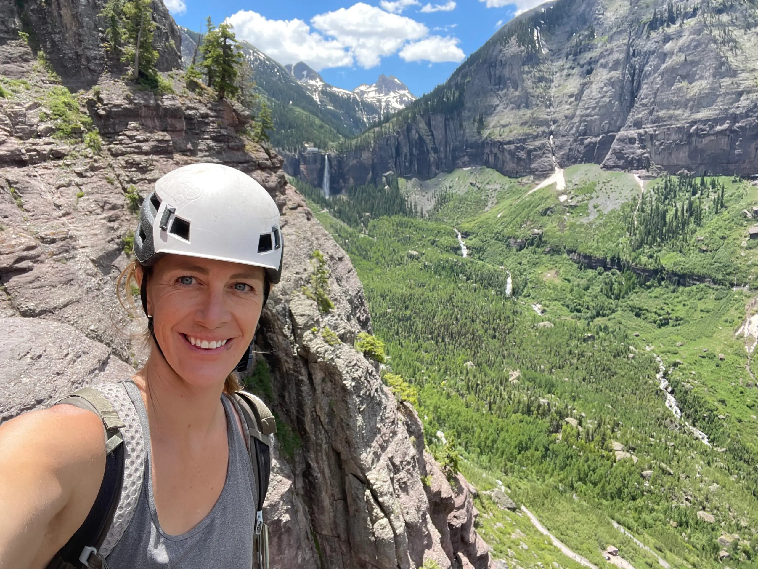 A person wearing a helmet and backpack smiles while standing on a rocky ledge with a scenic valley and distant mountains in the background in Denver, Colorado