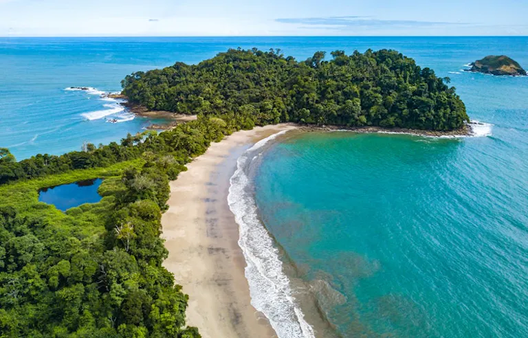 Aerial view of a sandy beach curving along turquoise water, bordered by dense green forest and a small island in the distance.