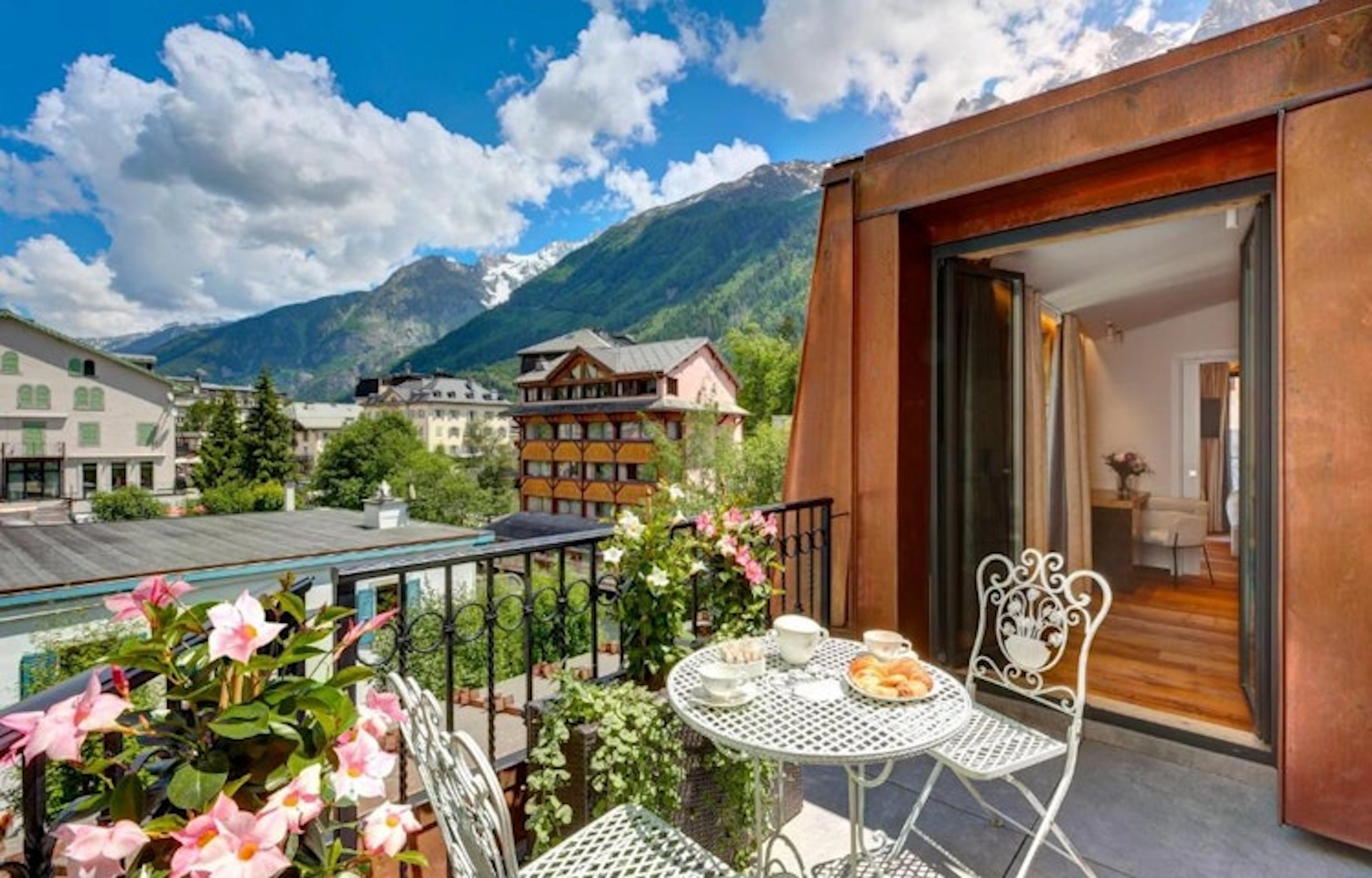 A balcony with a wrought iron table and chairs, set with breakfast items, overlooks a picturesque mountain village with lush greenery and clear blue skies in Chamonix, Europe, near the Alps