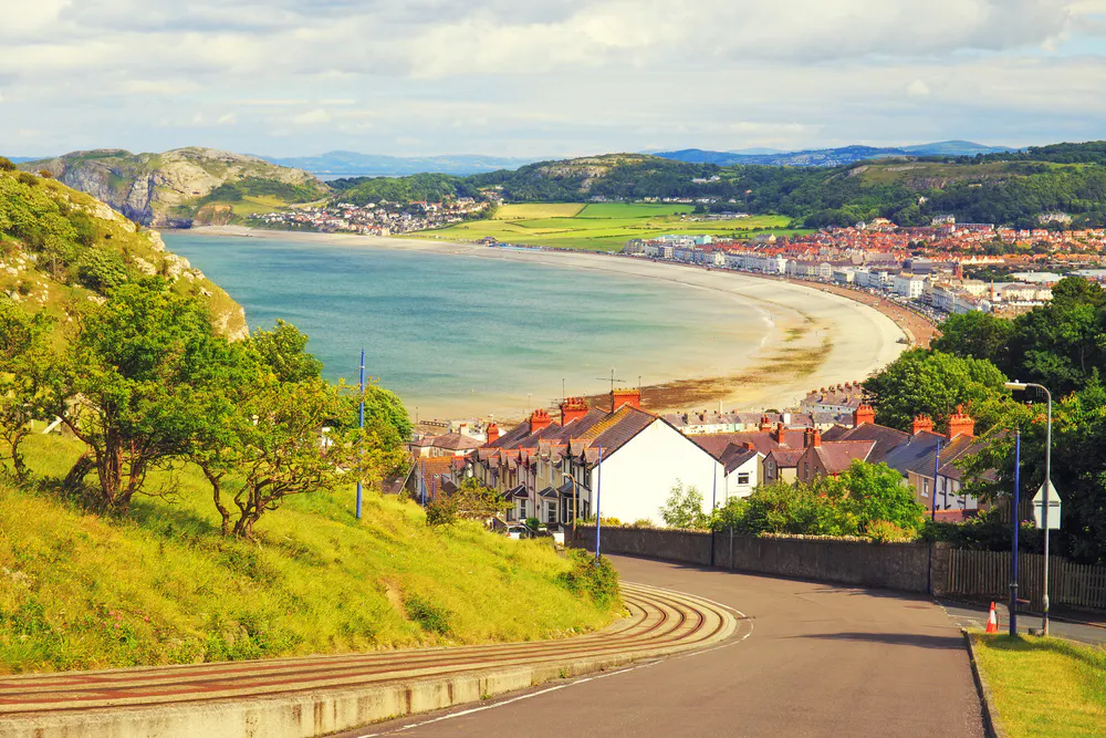 A scenic coastal town features curved roads, residential houses, and a long sandy beach with hills and greenery in the background on a partly cloudy day in England, Lake District.