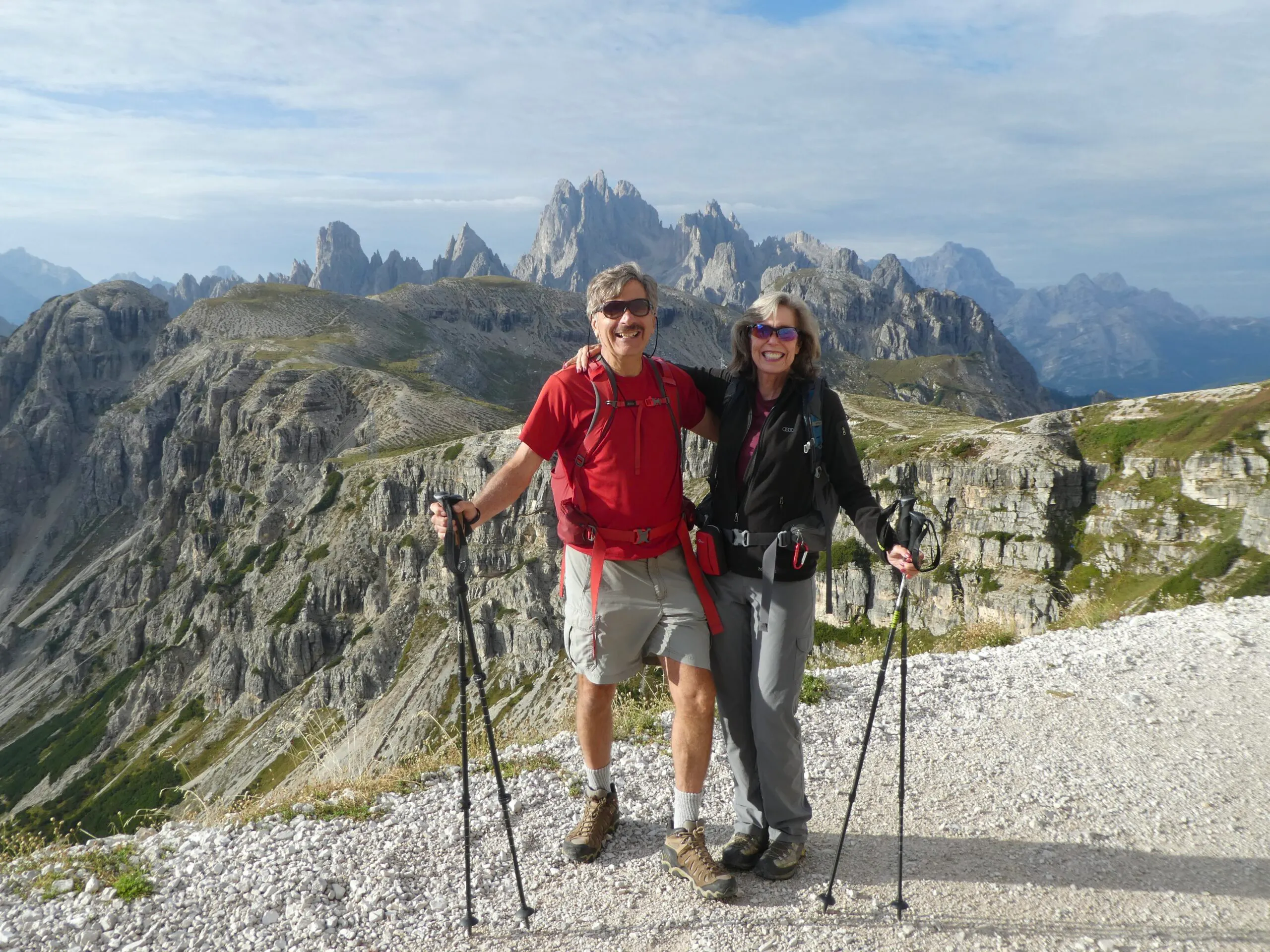 Two active hikers a couple with trekking poles stand on a rocky path in front of steep Dolomites mountain peaks under a partly cloudy sky in Italy, Europe