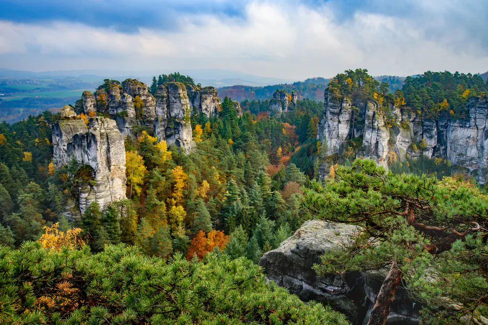 View of a scenic landscape featuring tall rock formations surrounded by a dense forest with autumn foliage under a cloudy sky in Bohemian Paradise