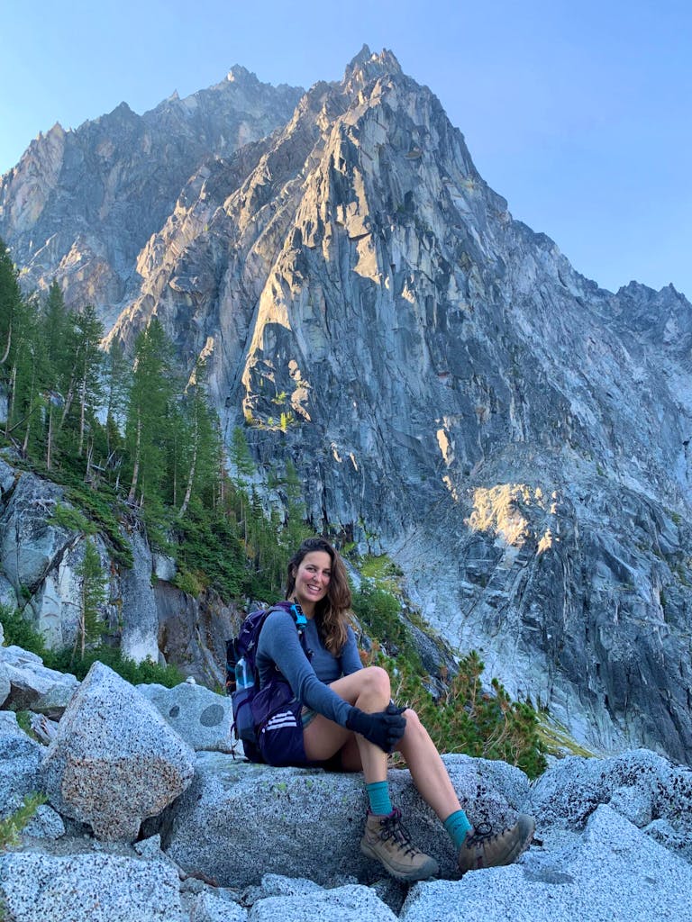 A person with long hair and a backpack is sitting on rocks with a mountainous backdrop in the pacific northwest. They are wearing hiking gear and are smiling at the camera.