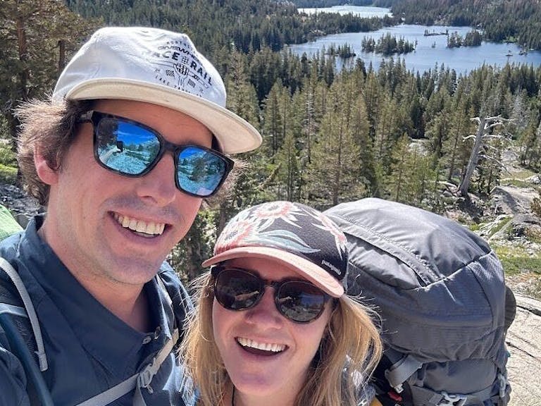 Two people wearing hats and sunglasses smile for a selfie during a hike. A lake and forested mountains are visible in the background.