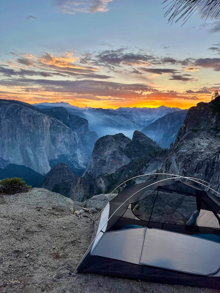 A tent is set up on a rocky ledge overlooking a mountainous landscape at sunset, with the sky displaying vibrant colors.