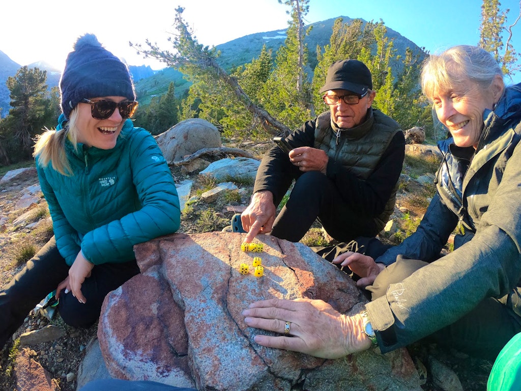 Three people sitting outdoors on rocky terrain, playing a game with yellow dice on a large rock. Mountain and trees visible in the background.