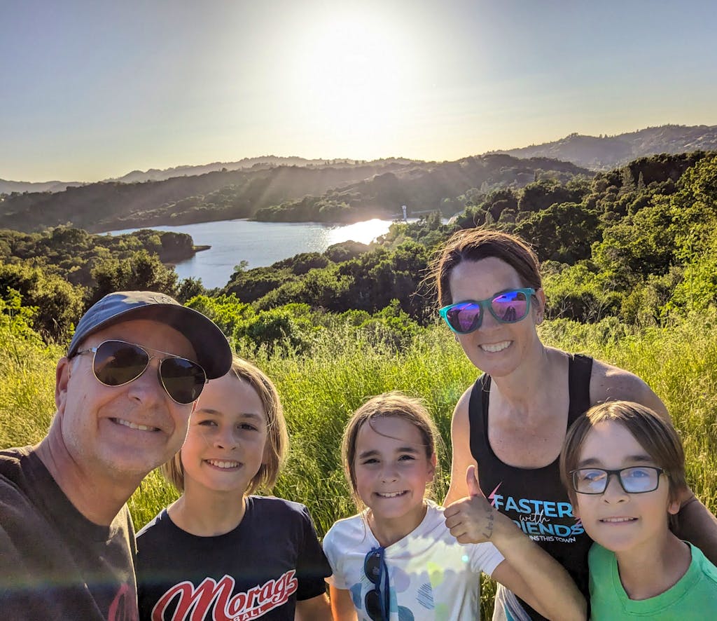 A group of four people, including two adults and two children, are smiling and posing for a photo outdoors with a scenic, sunlit landscape of hills and water in the background in Lafayette Reservoir, California