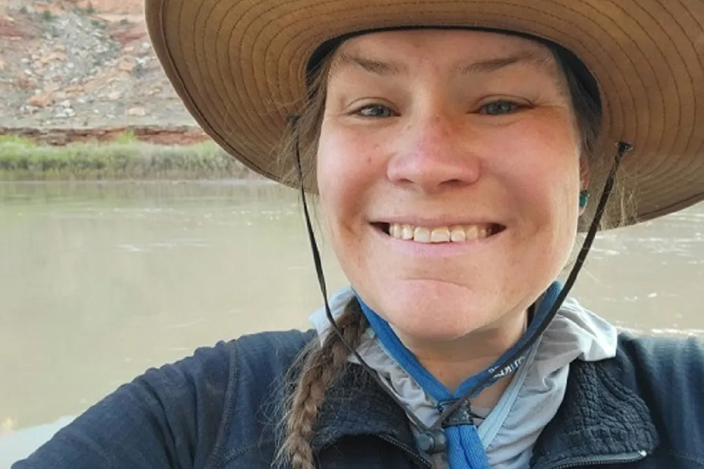 Person wearing a wide-brimmed hat and jacket smiles at the camera by a river, with rocky terrain and greenery in the background.