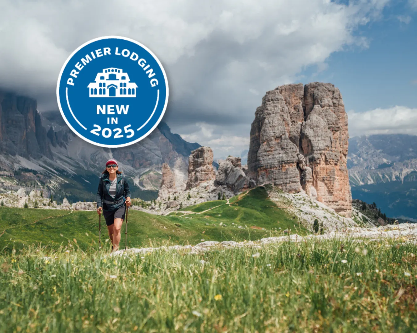 A hiker walks on a grassy trail in a mountainous landscape with rock formations. A badge overlay reads, "Premier Lodging New in 2025.