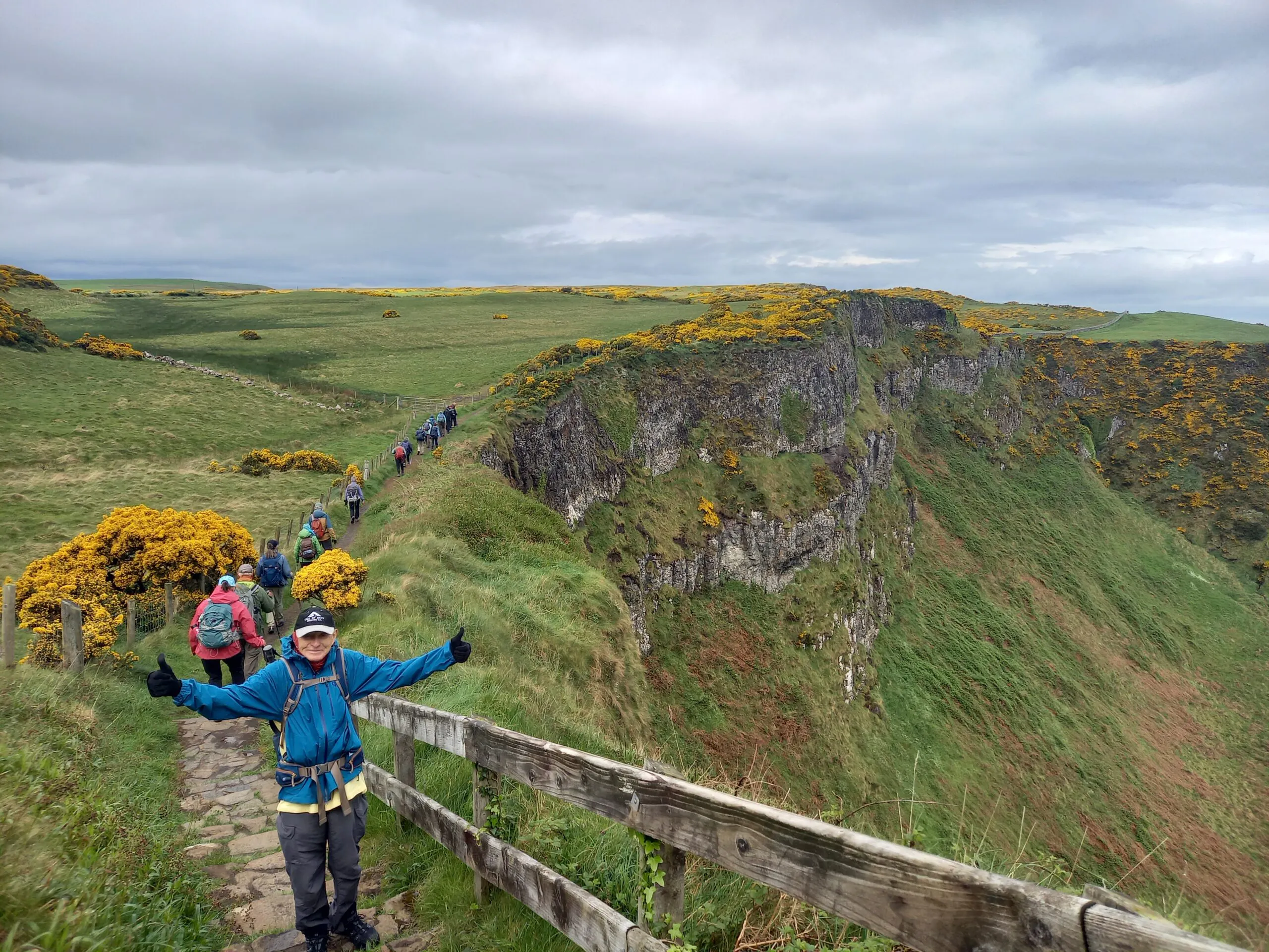 A group of hikers walks along a stone path beside a steep grassy cliff, with one person in the foreground posing with arms outstretched.