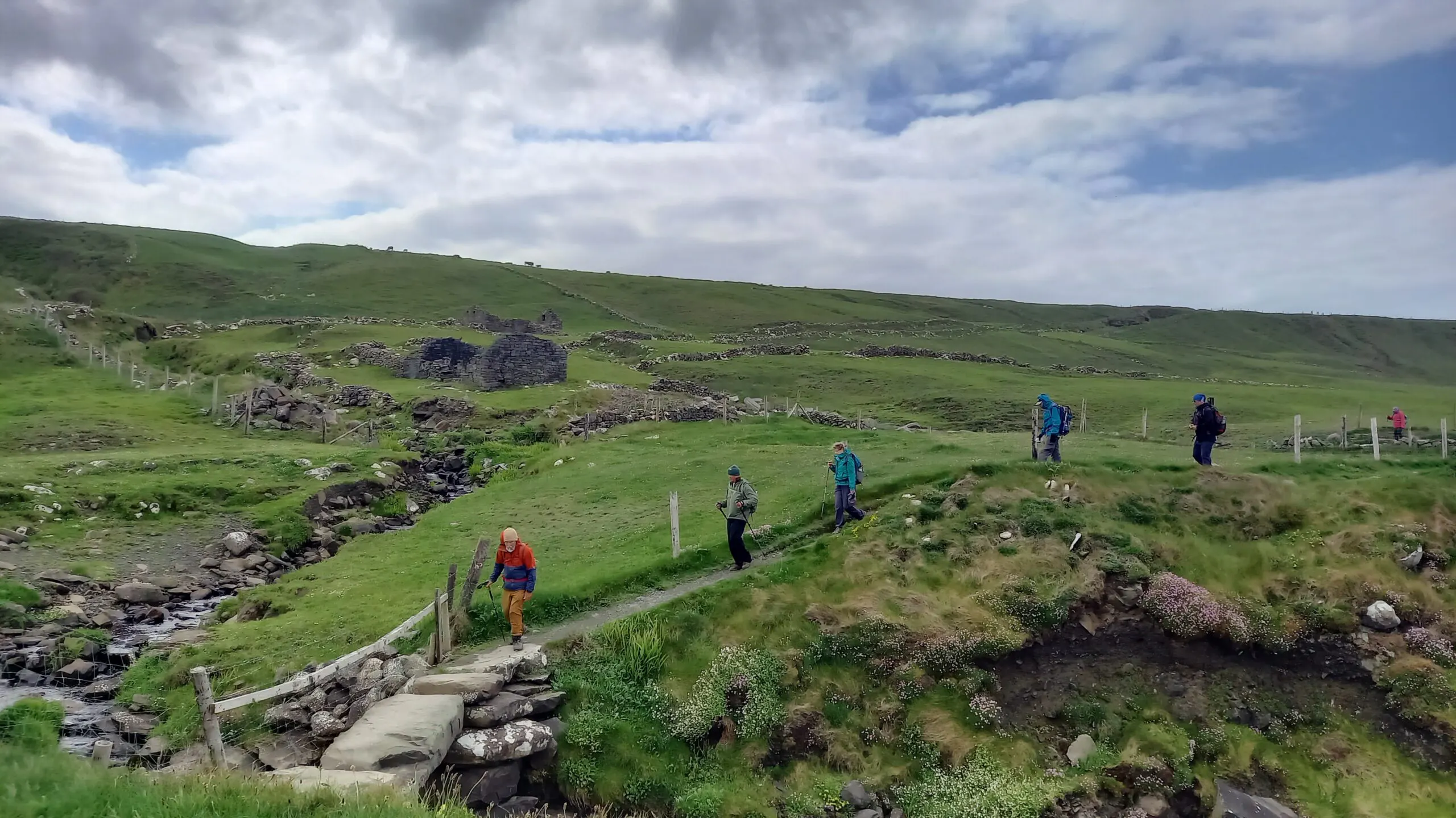 Several people hike along a narrow path in a green, hilly landscape with scattered rocks and a cloudy sky overhead.