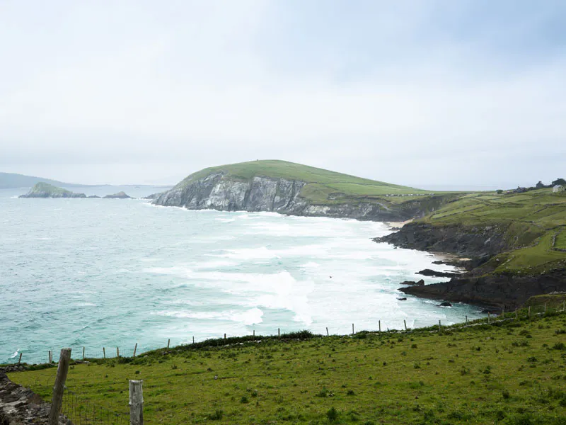 Coastal landscape with grassy hills, rocky cliffs, and waves crashing against the shoreline under a cloudy sky.