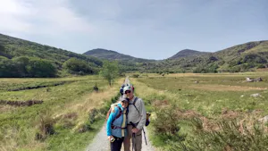 Two people wearing hiking gear stand together on a trail surrounded by grassy hills and mountains under a clear sky.