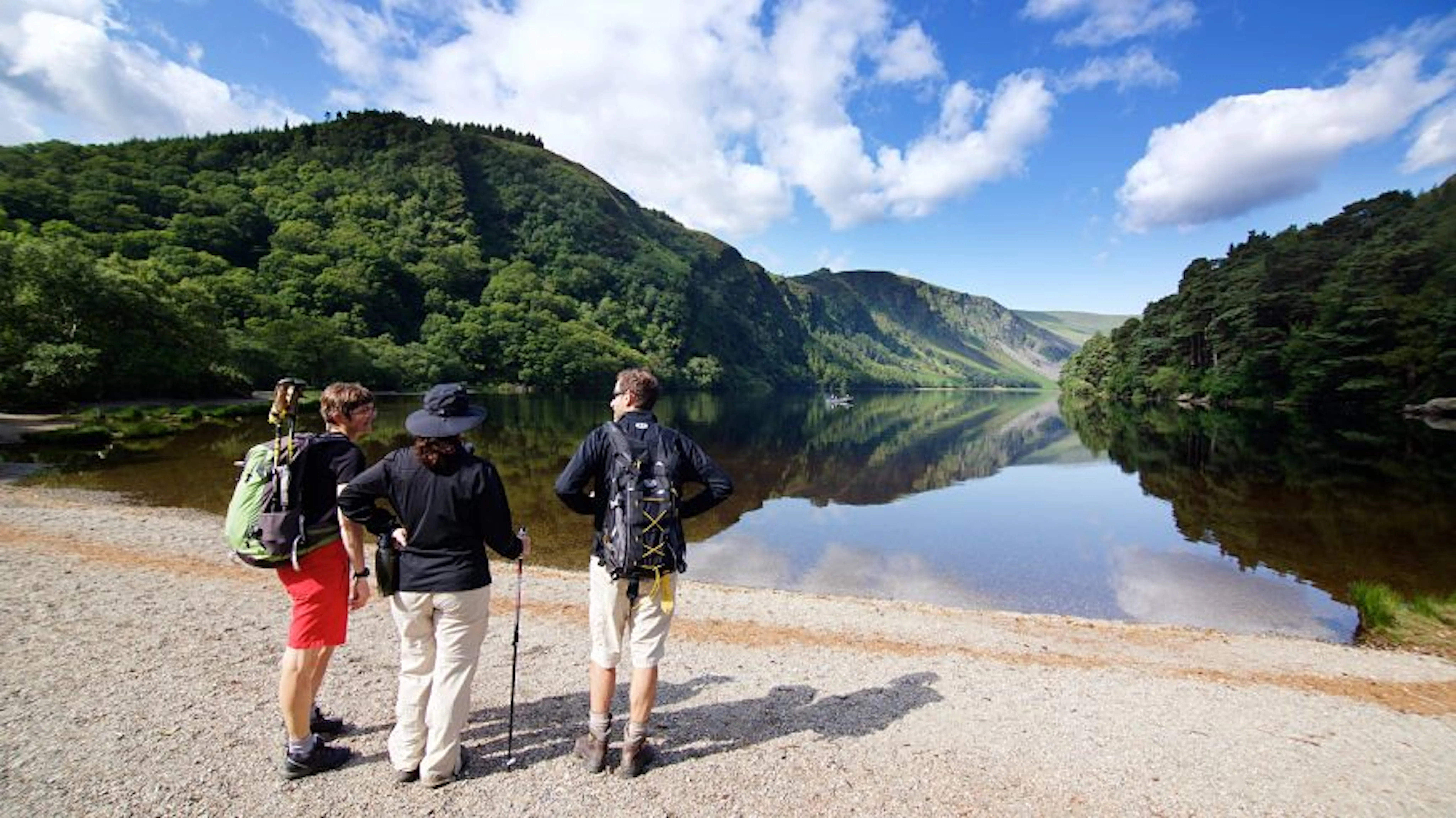 Three hikers stand on a gravel path by a calm lake, looking at forested hills and blue sky with clouds reflected in the water.