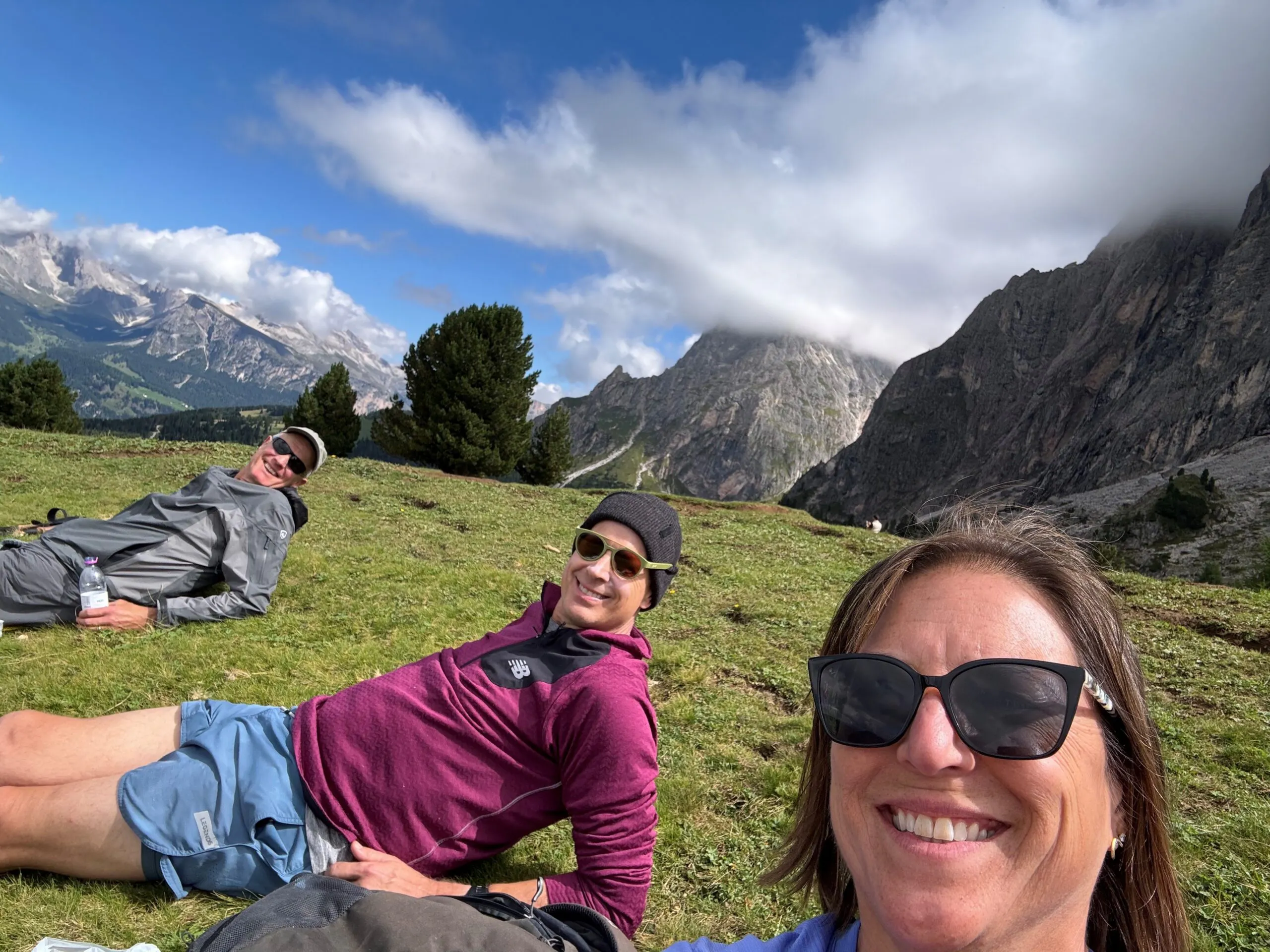 Three people wearing sunglasses relax on a grassy hillside with mountains, trees, and clouds in the background.