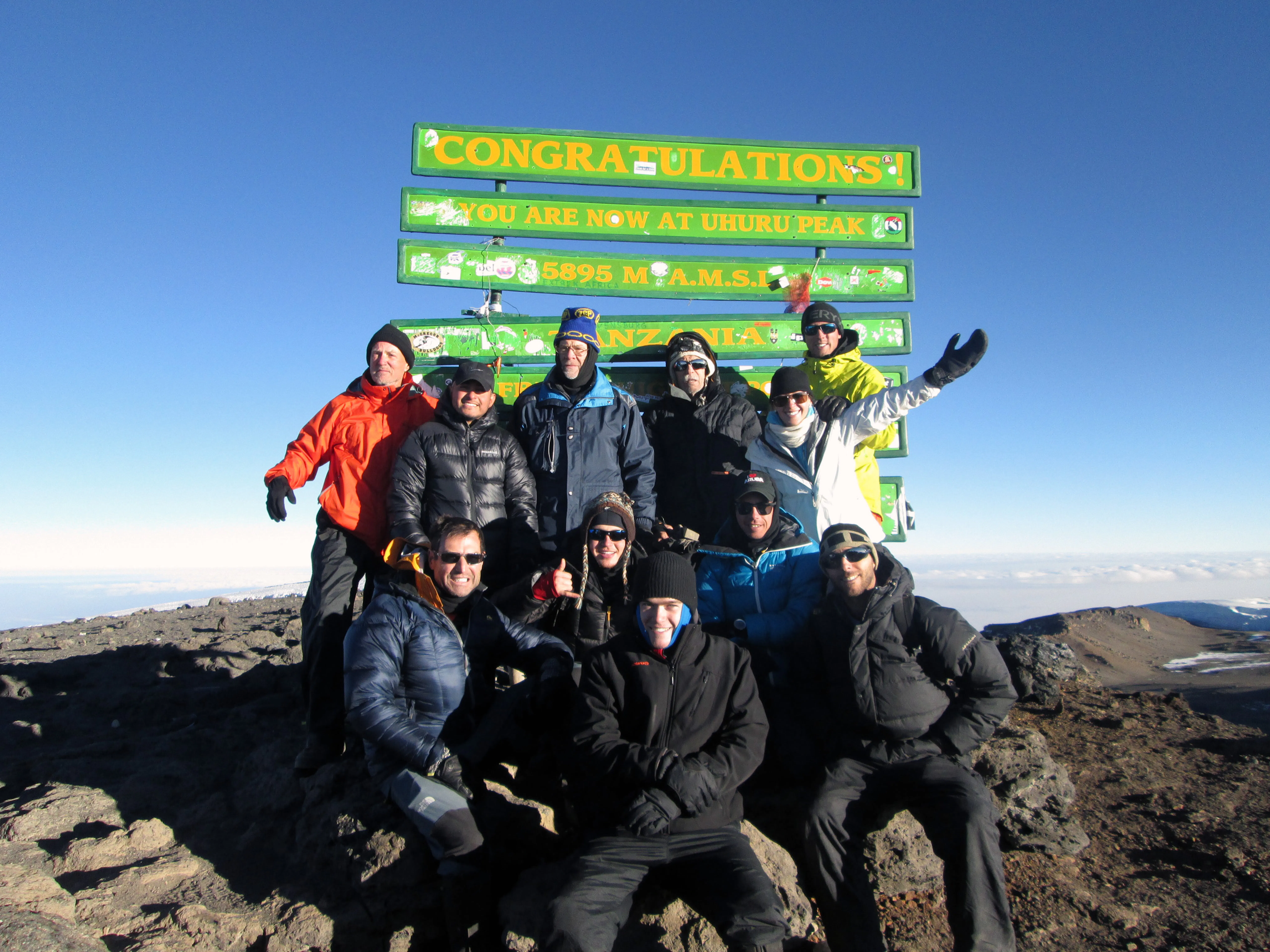A group of people in winter clothing pose and smile after hiking to Uhuru Peak, the summit of Mount Kilimanjaro, under a clear sky.