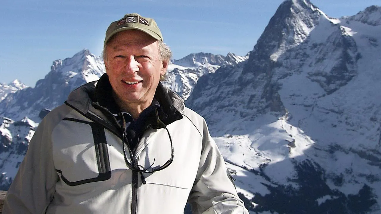 A man wearing a light jacket and cap stands outdoors with snow-covered mountains in the background under a clear sky.