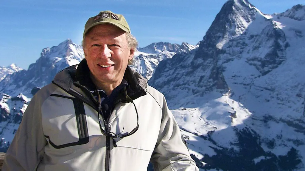 A man wearing a light jacket and cap stands outdoors with snow-covered mountains in the background under a clear sky.