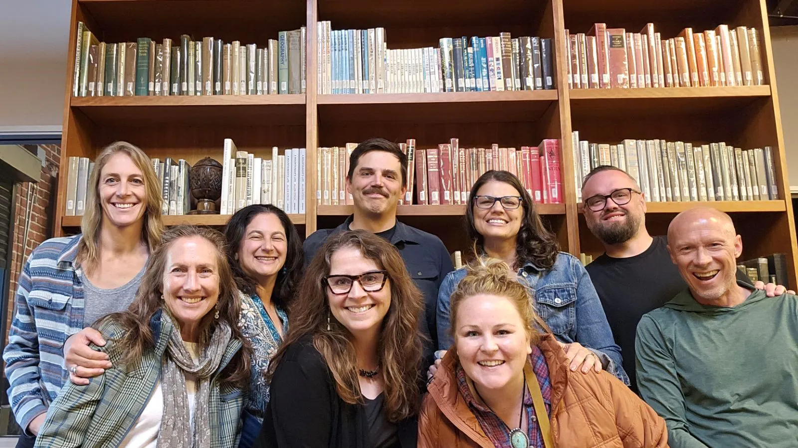 A group of ten adults poses and smiles in front of shelves filled with books in a library or study room, perhaps planning their next custom travel adventure together.