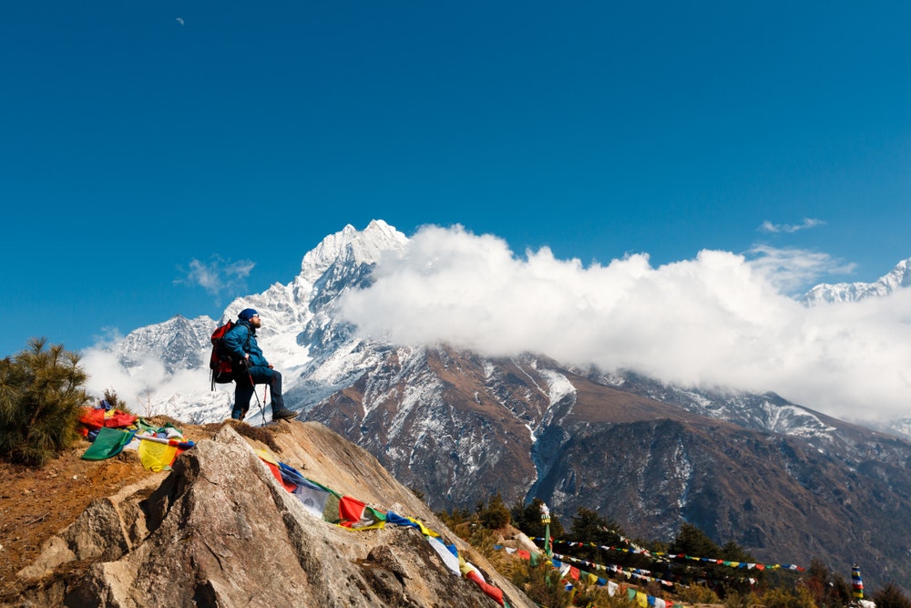 Everest Base Camp Trek. A man stands on a hill and looks at the horizon. In the background are mountains and clouds. In the frame Nepalese prayer flags with prayers.