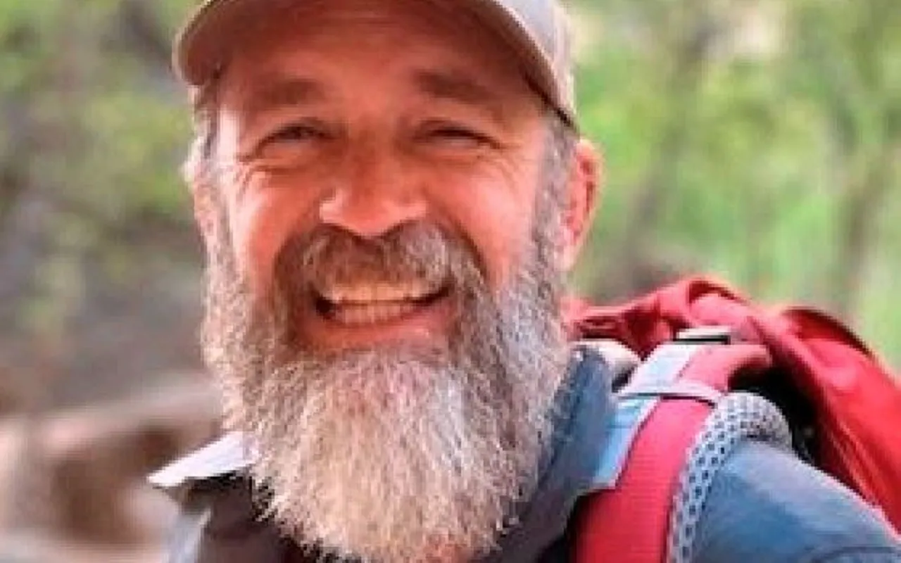 A bearded man wearing a cap and backpack smiles outdoors with blurred greenery in the background.