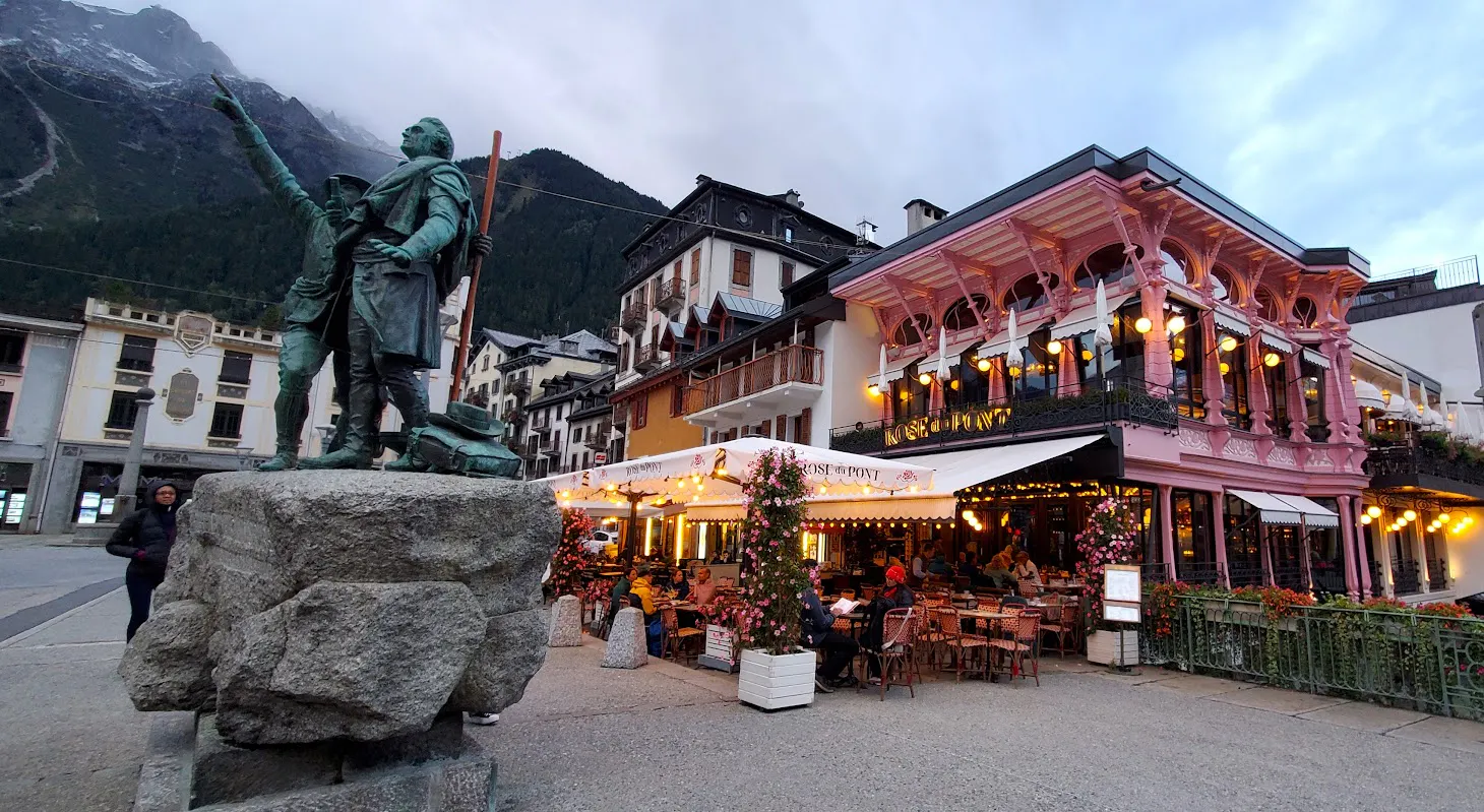A bronze statue stands on a stone pedestal near a busy outdoor restaurant with pink trim, surrounded by alpine buildings and mountains—perfect for those enjoying alp hiking or exploring chamonix and zermatt hiking trails.