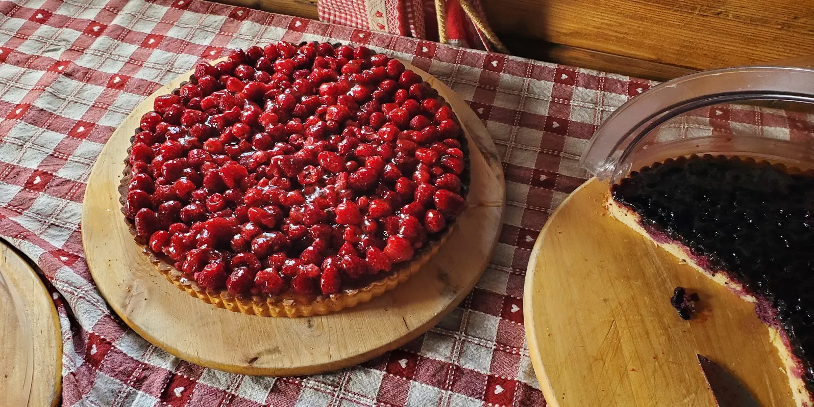 A raspberry tart and a partially sliced blueberry tart on wooden boards, placed on a red and white checkered tablecloth—perfect treats after a day of Chamonix and Zermatt hiking in the Alps.