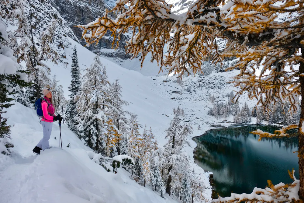 A person in winter gear stands on a snowy trail, overlooking a snow-covered forest and a partially frozen lake surrounded by mountains, ready for a Banff adventure.