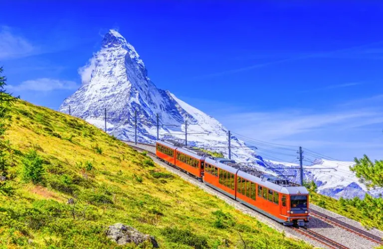 An orange train travels uphill on a railway with the snow-covered Matterhorn mountain in the background, capturing the essence of alp hiking adventures under a clear blue sky.