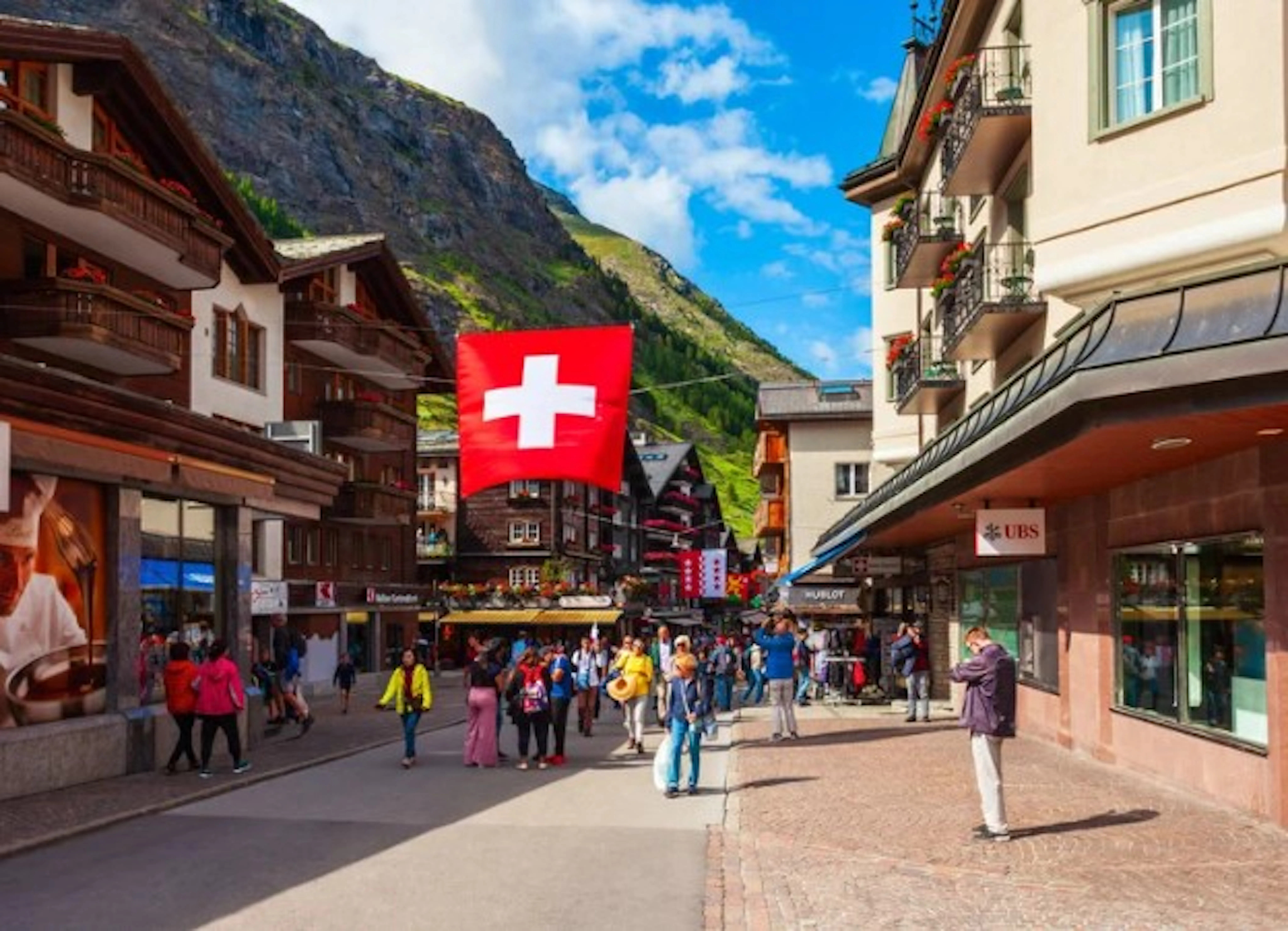 A busy street scene in a Swiss mountain town, featuring people walking, shops, buildings with balconies, and a large Swiss flag hanging overhead—an inviting spot for alps hiking enthusiasts to explore after their adventures.
