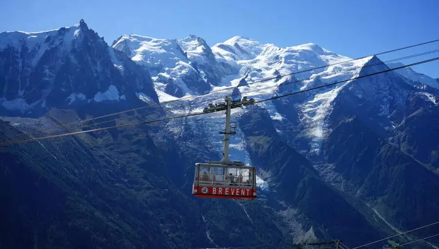 A red cable car labeled "BREVENT" glides in front of snowy mountain peaks under a clear blue sky, offering stunning views for those enjoying alps hiking or planning adventures between Chamonix and Zermatt.