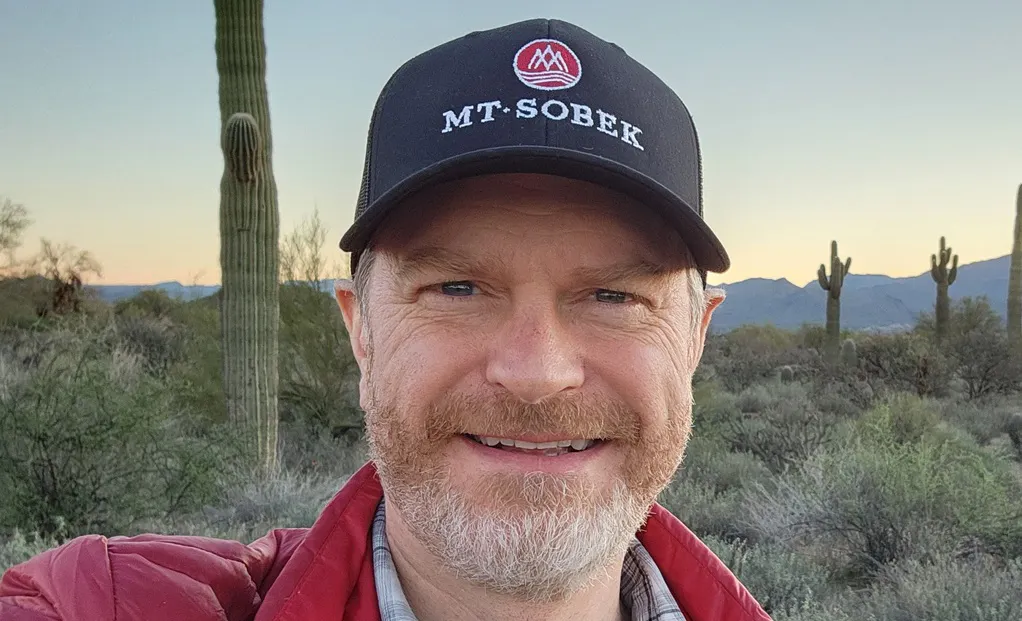 A man with a beard wearing a black "Mt. Sobek" cap and red jacket smiles outdoors in a desert landscape with cacti and mountains in the background.