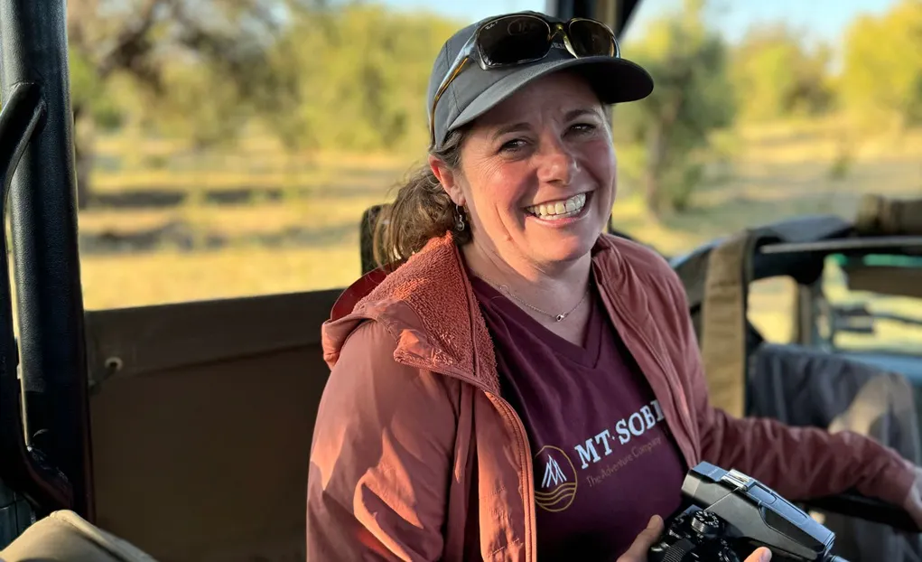 Woman smiling in an outdoor vehicle, wearing a cap, sunglasses, and a maroon “Mt. Sobek” shirt, holding a camera; trees and sunlight in the background.