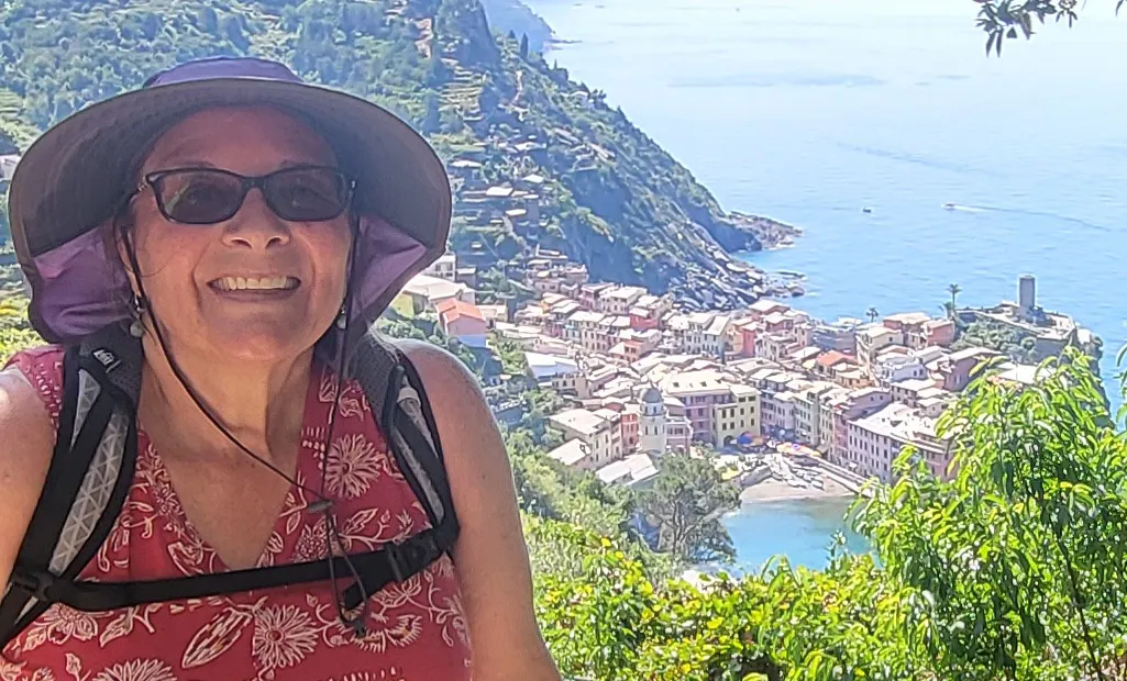 Woman wearing a sun hat and sunglasses smiles in the foreground, with a coastal village and blue sea visible below in the background.