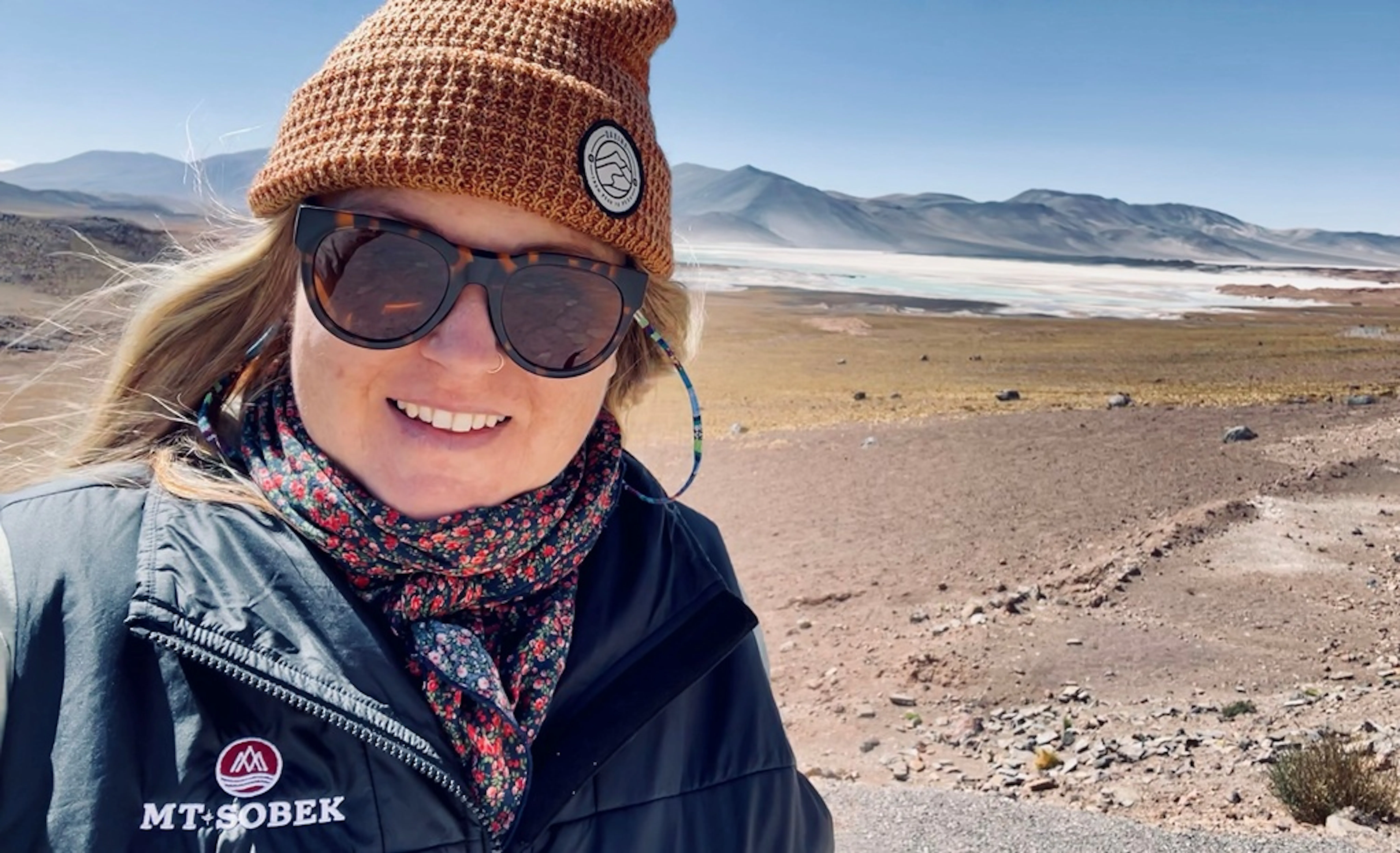 Woman wearing sunglasses and a beanie stands outdoors in a dry, mountainous landscape with a salt flat visible in the background.