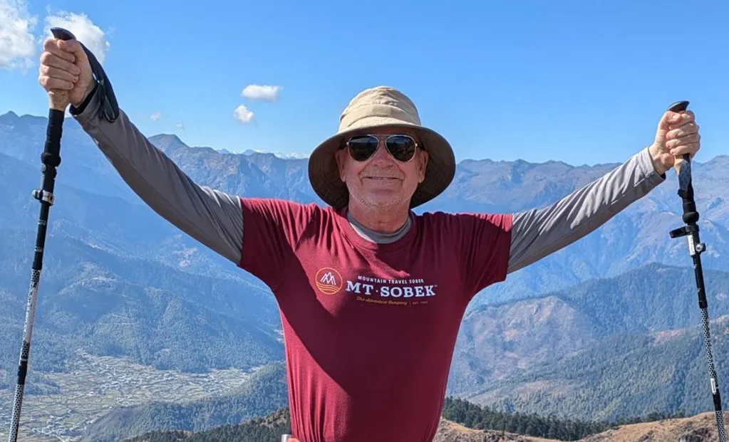 Man wearing a hat and sunglasses stands outdoors with hiking poles raised, mountains and blue sky in the background.