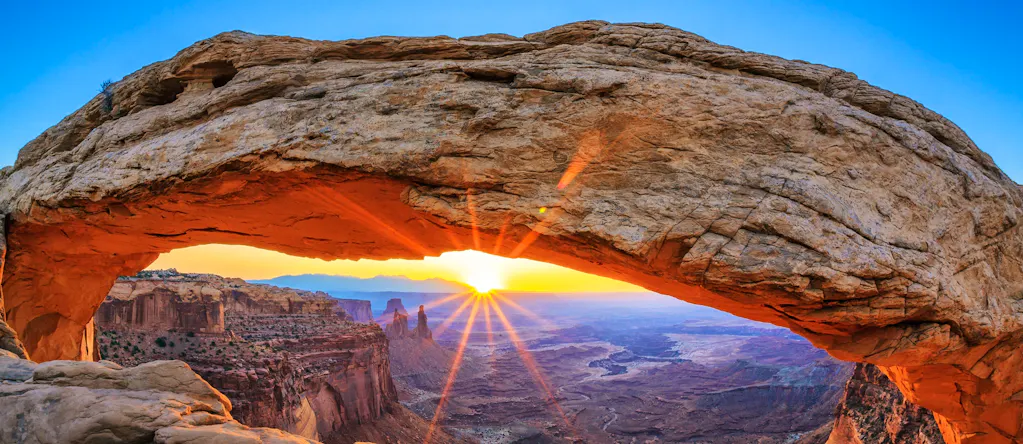 Sunrise shines through the Mesa Arch in Utah National Parks, revealing a canyon landscape below, perfect for hiking under a clear blue sky.