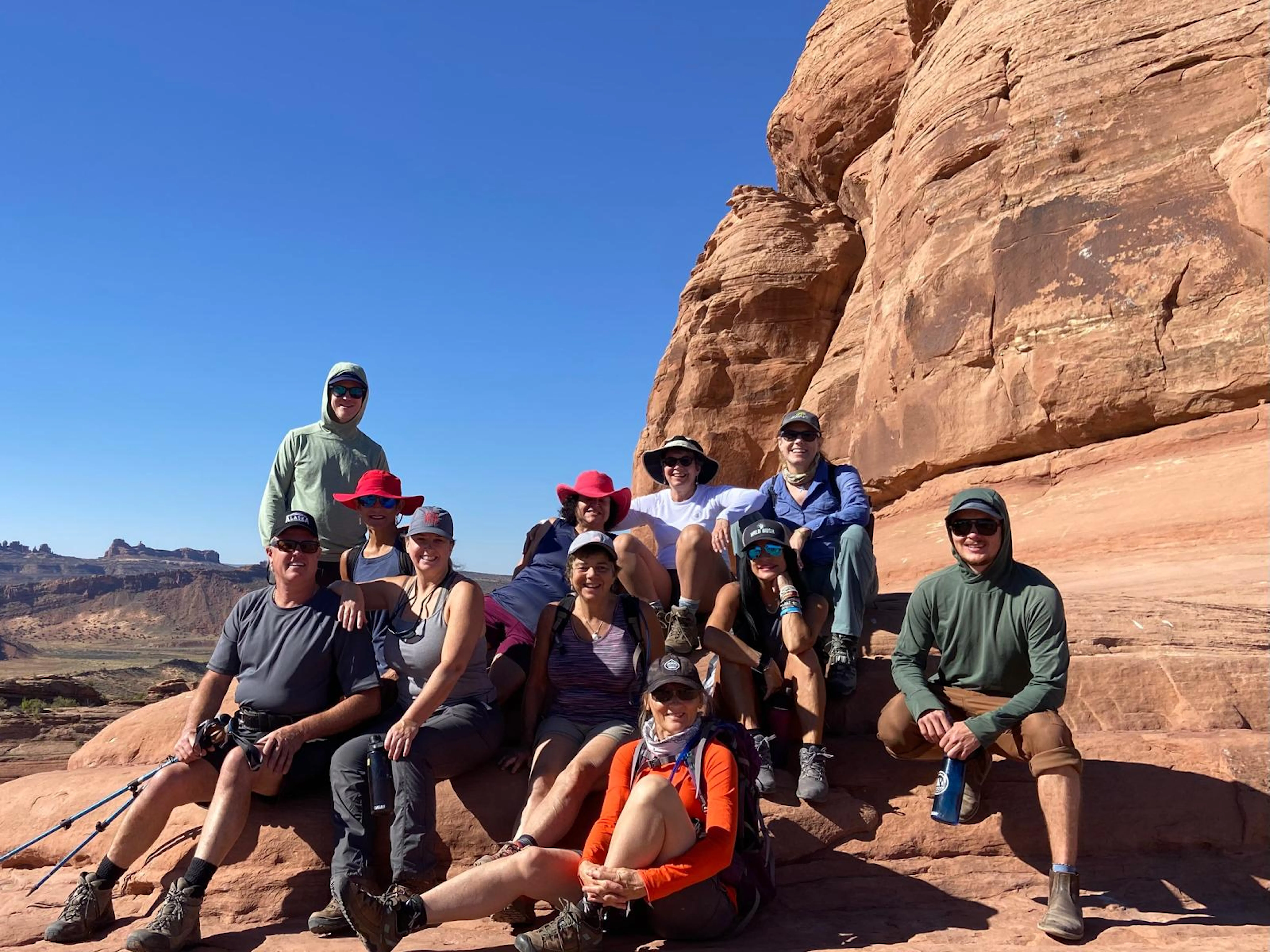 A group of people in hiking attire sit and pose together on red rock formations under a clear blue sky, capturing the adventurous spirit of Utah national parks hiking.