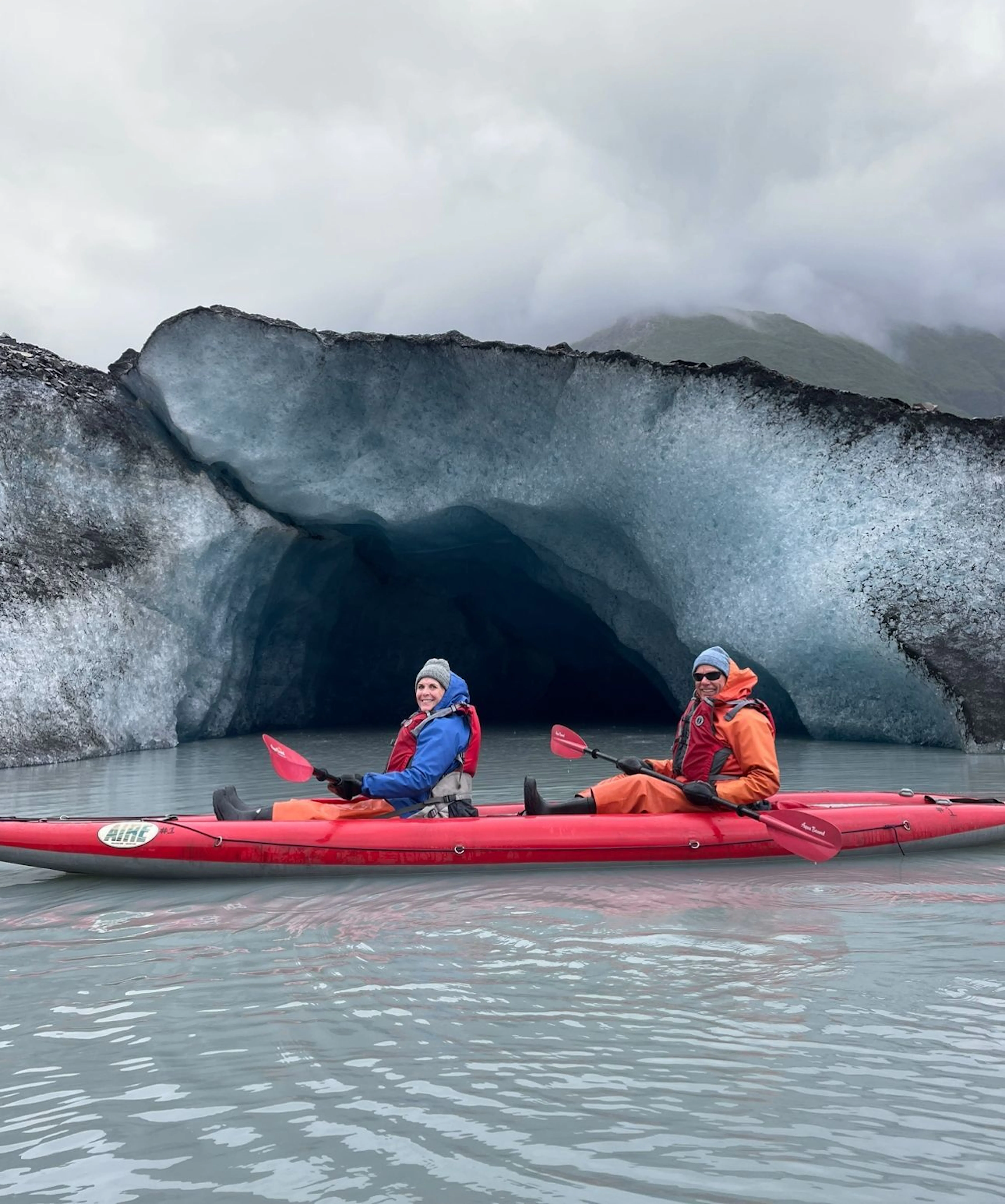 Two people in a red kayak paddle on glacial water before a blue ice cave, dressed in warm jackets and beanies under a cloudy sky—an unforgettable moment from Alaska tours.