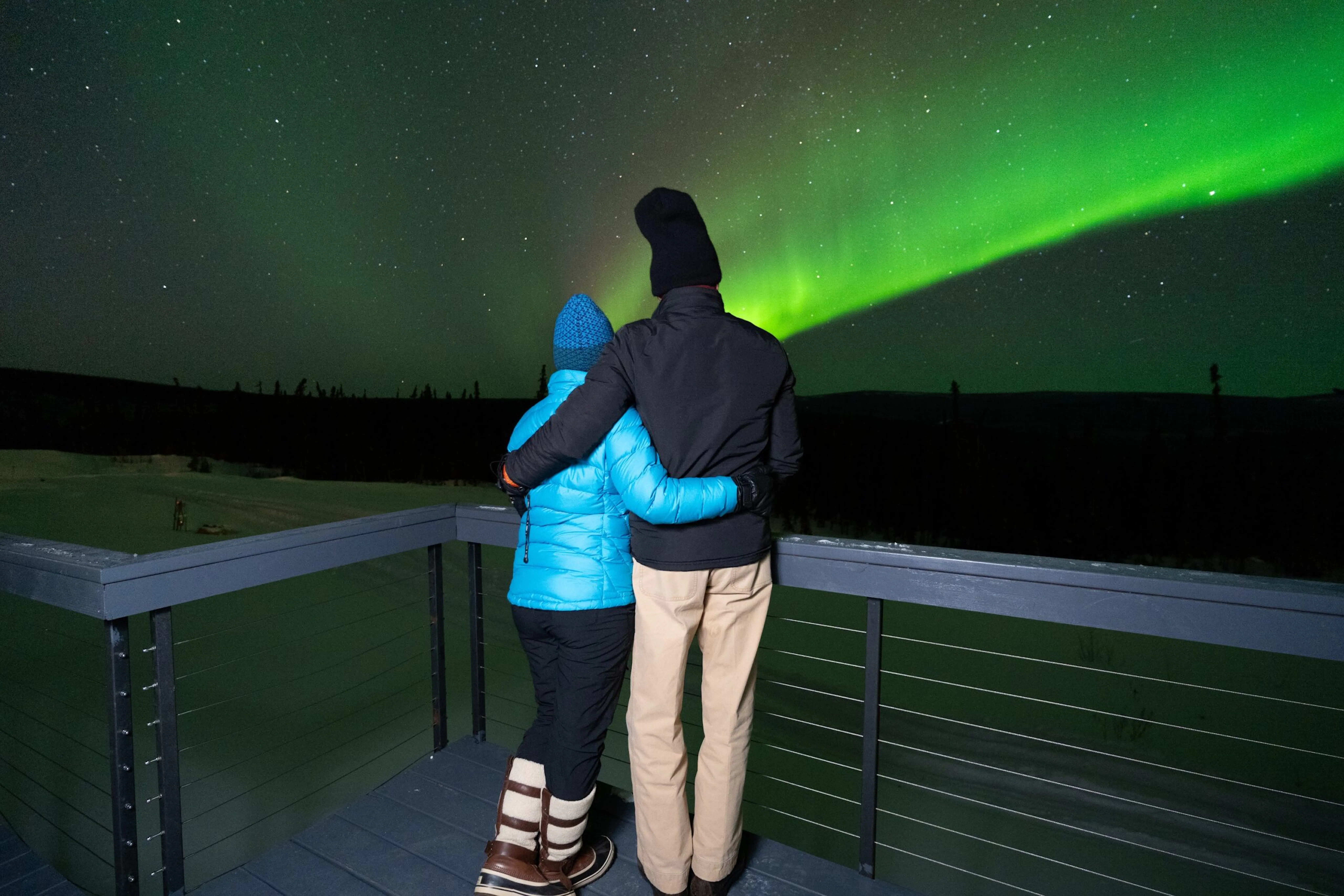 Two people in winter clothing stand on a deck at night, watching a bright green aurora borealis display in the sky—an unforgettable moment during one of their Alaska tours.